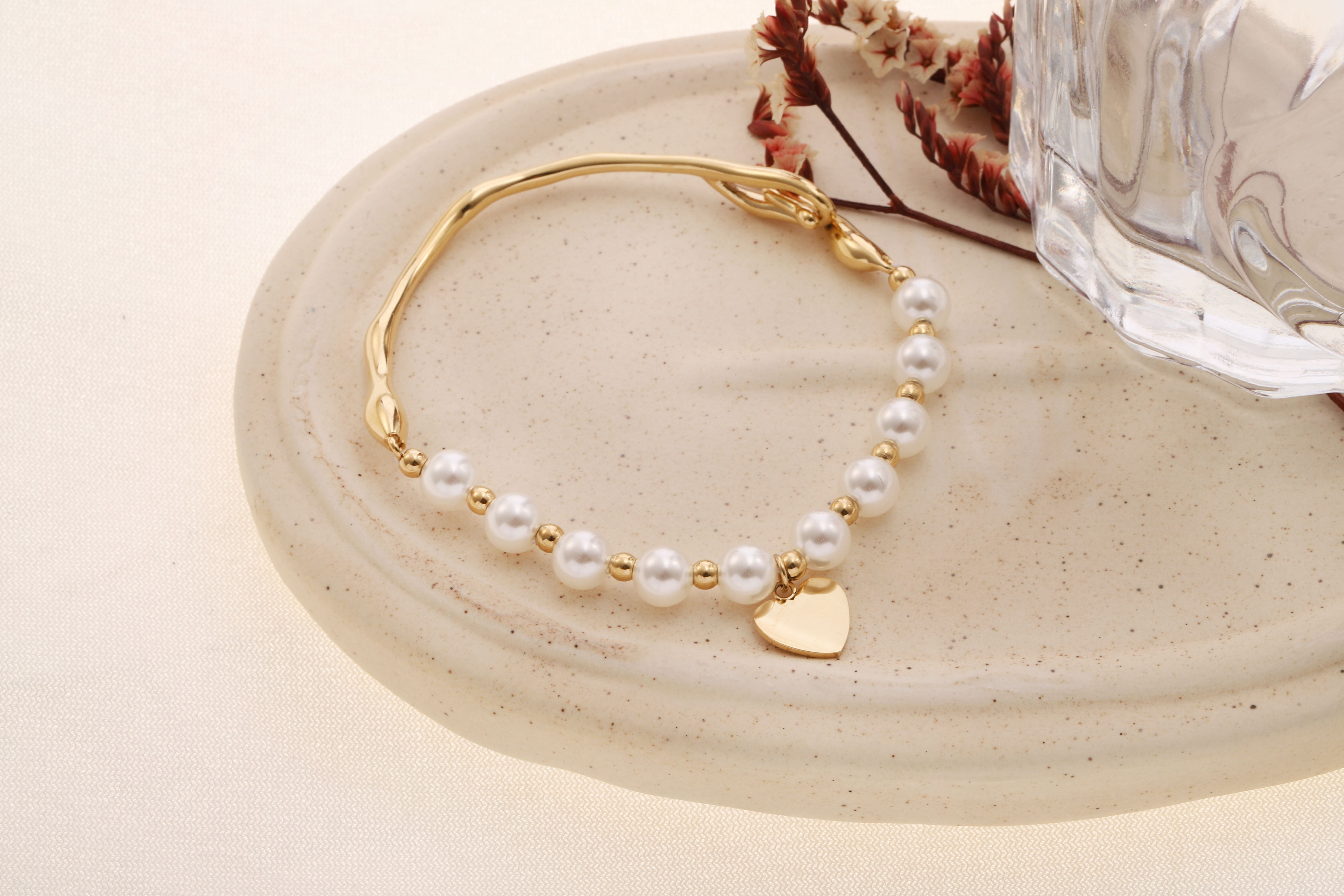 Bracciale semirigido in oro con perla bianca laterale e ciondolo a forma di cuore, chiusura a gancio