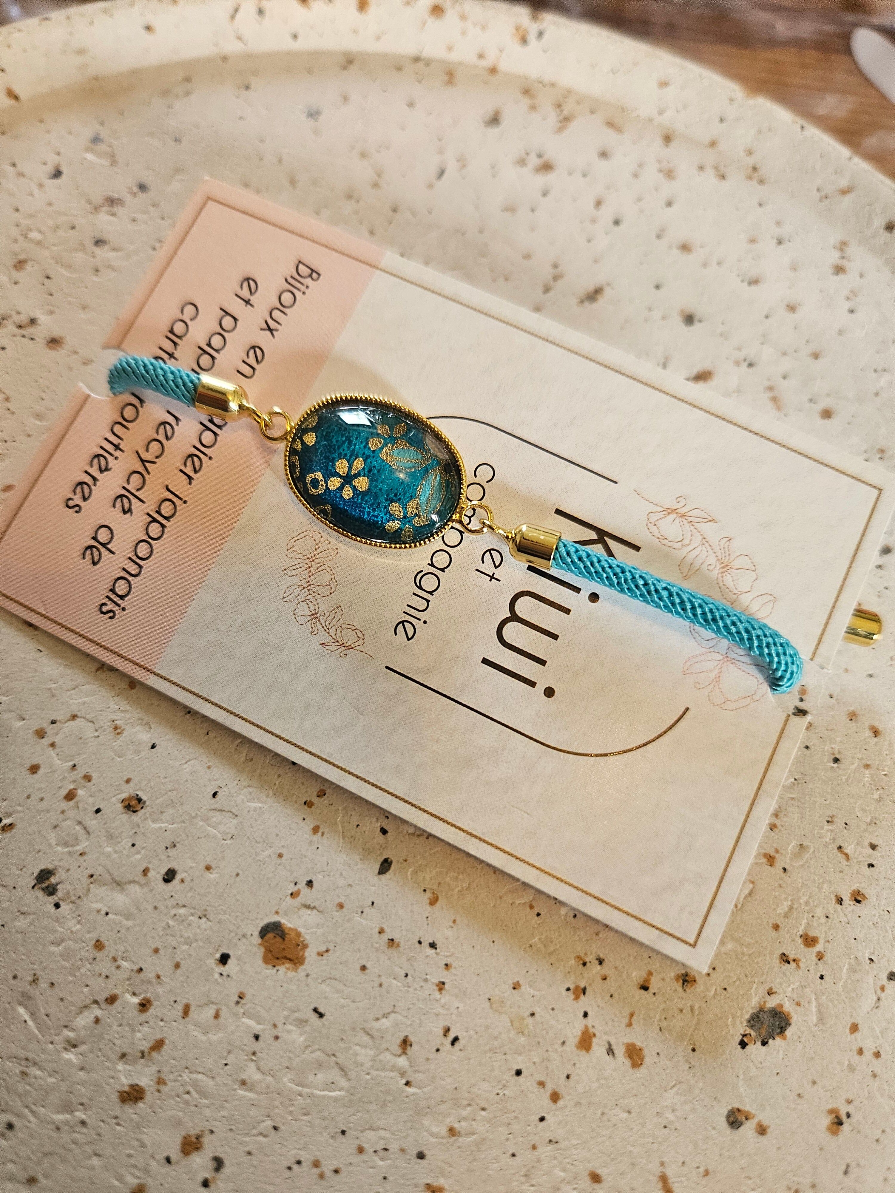 Adjustable blue cotton bracelet with a blue cabochon, oval charm, and gold flower.